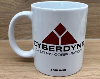 May include: A white ceramic mug featuring a dark red triangular logo above the black text CYBERDYNE SYSTEMS CORPORATION. Below this, the text FLEXI ARCADE is printed in small black letters with a small red icon to the left of the word.