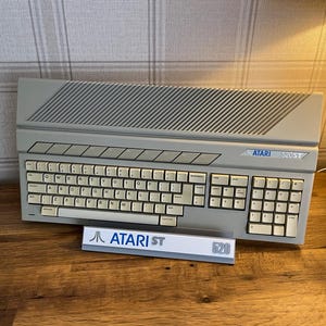 May include: A beige Atari 520ST home computer with a QWERTY keyboard, a numeric keypad, and a series of function keys. The unit features a slanted, vented top panel and a small display stand in front with the Atari ST logo and 520 text printed.
