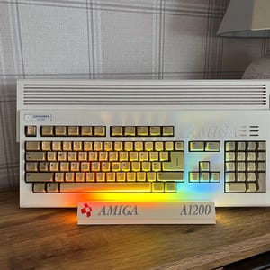 May include: A beige Commodore Amiga 1200 computer with a QWERTY keyboard and a horizontal vent pattern on the top casing. A white stand in front displays the red Amiga logo and the text A1200, illuminated by a vibrant rainbow light effect.