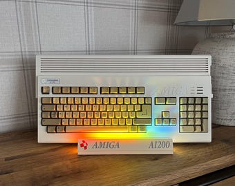 May include: A beige Commodore Amiga 1200 computer with a QWERTY keyboard and a horizontal vent pattern on the top casing. A white stand in front displays the red Amiga logo and the text A1200, illuminated by a vibrant rainbow light effect.