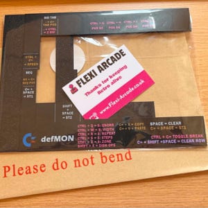 Könnte beinhalten: Eine schwarze und transparente Kunststofffolie mit Tastenkombinationen, eine weiße Visitenkarte mit dem Text "FLEXI ARCADE" und "Thanks for keeping Retro alive" und ein brauner Umschlag mit dem Text "Please do not bend".