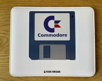 May include: A rectangular white mouse pad featuring a centered graphic of a dark blue 3.5-inch floppy disk. The disk label displays the blue and red Commodore logo with the text Commodore below it. The text FLEXI ARCADE is printed at the bottom.