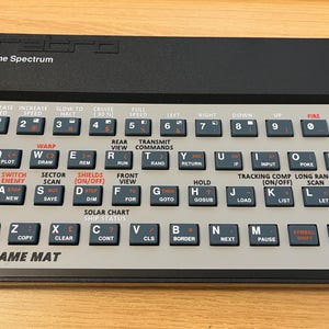 Op de afbeelding: Een grijs en zwart toetsenbord met een verscheidenheid aan toetsen die zijn gelabeld met letters, cijfers en commando's. Het toetsenbord heeft een zwarte rand en de tekst "The Spectrum" en "CODENAME MAT" erop gedrukt.