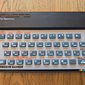 Könnte beinhalten: Eine Retro-Computer-Tastatur in Schwarz und Grau mit blauen Tasten und weißer Beschriftung. Die Tastatur trägt oben den Text "retro The Spectrum" und unten "AIRBORNE RANGER". Die Tasten sind mit Buchstaben, Zahlen und Befehlen beschriftet.