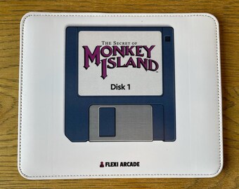 May include: A rectangular white mouse pad featuring a graphic of a dark blue floppy disk. The disk label reads The Secret of Monkey Island Disk 1 in purple and black text. The bottom center displays the text Flexi Arcade with a small icon.