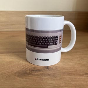 Peut inclure: Un mug en céramique blanche orné d'une impression graphique représentant un clavier d'ordinateur vintage avec des touches sombres et un boîtier gris clair. Sous le clavier, le texte FLEXI ARCADE est imprimé en noir avec une petite icône de joystick rouge.