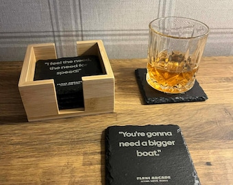 May include: A set of slate coasters with quotes from movies. One coaster reads "You're gonna need a bigger boat." Another says "I feel the need, the need for speed!" The coasters are in a wooden holder.