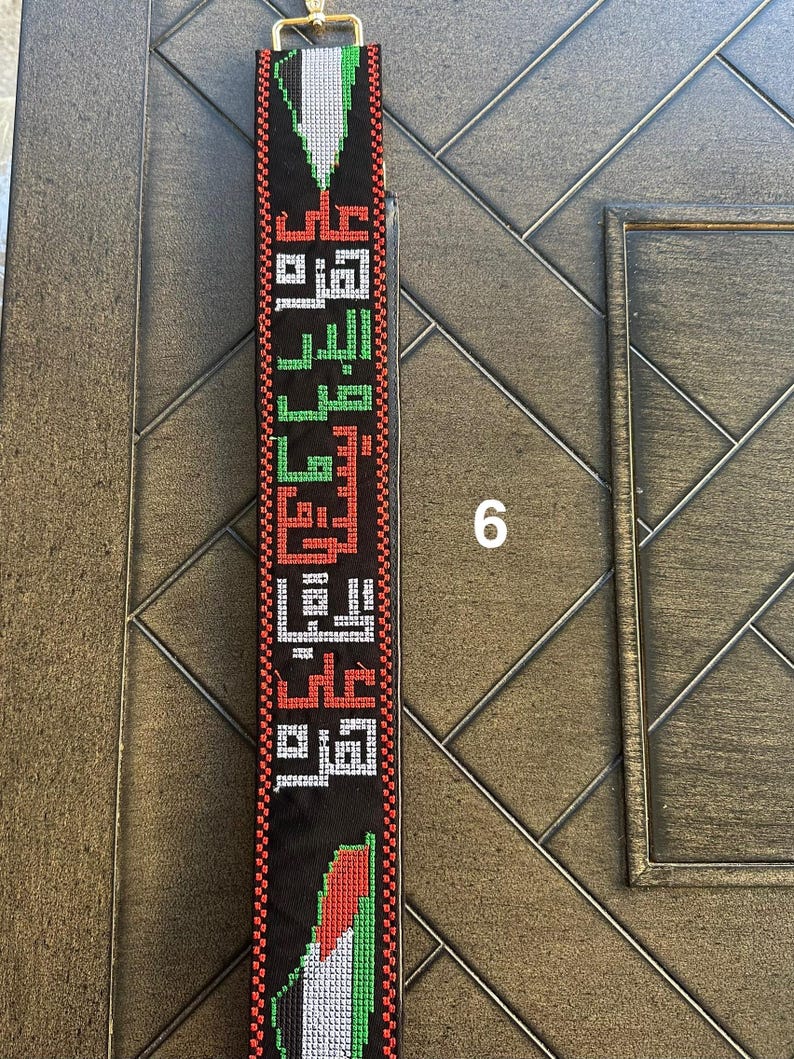 May include: A black fabric strap with a woven design featuring the Palestinian flag colors of red, green, white, and black. The strap has Arabic text woven into the design.
