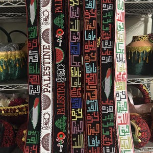 May include: Six colorful woven belts with the word "Palestine" written in Arabic script. The belts are decorated with various symbols, including a watermelon, a mosque, and a key.