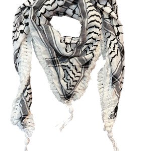 May include: A white and black keffiyeh scarf with a geometric pattern and fringed edges. The scarf has a traditional design with black lines and shapes on a white background. The scarf is made of a soft material and has tassels at the ends.