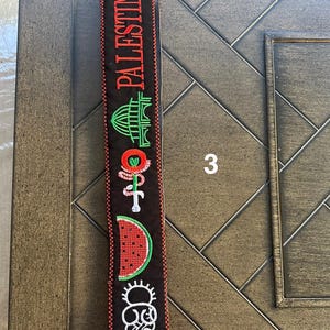 May include: Black and red fabric strap with embroidered images of a building, a watermelon, a flower, and a person. The strap has the word "PALESTINE" embroidered on it in red.