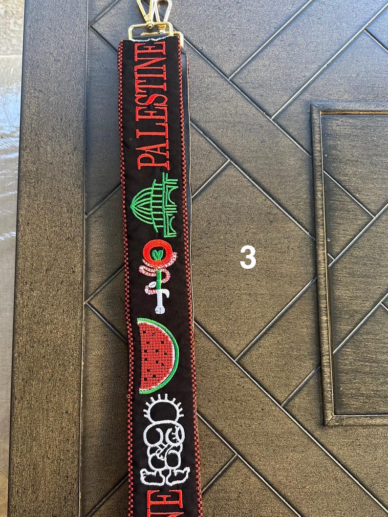 May include: Black and red fabric strap with embroidered images of a building, a watermelon, a flower, and a person. The strap has the word "PALESTINE" embroidered on it in red.
