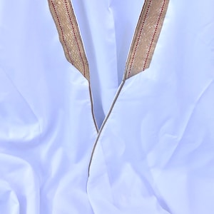 May include: White garment with a V-neck design. The neckline is trimmed with a decorative gold and red patterned border. The fabric appears to be lightweight and flowing, suggesting a robe or tunic.