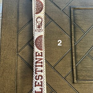 May include: A white fabric strap with brown embroidery. The embroidery includes the word "PALESTINE" and a stylized image of a sun and a crescent moon.