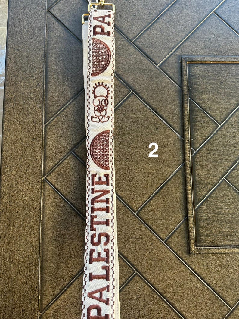 May include: A white fabric strap with brown embroidery. The embroidery includes the word "PALESTINE" and a stylized image of a sun and a crescent moon.