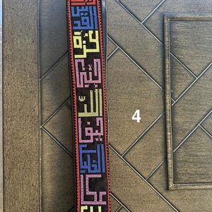 May include: A black fabric strap with colorful embroidered Arabic script. The strap has a gold metal clasp.