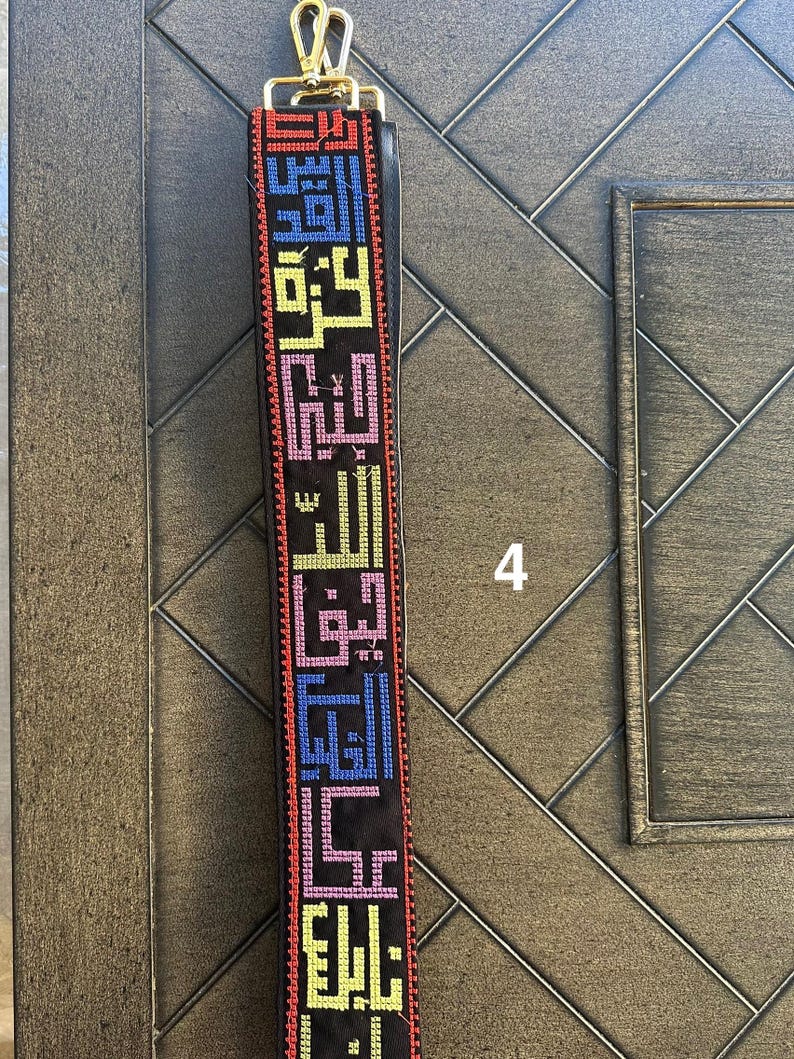 May include: A black fabric strap with colorful embroidered Arabic script. The strap has a gold metal clasp.