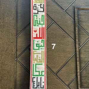 May include: A white strap with red, green, black, and gold embroidered Arabic text. The strap has a gold metal buckle.