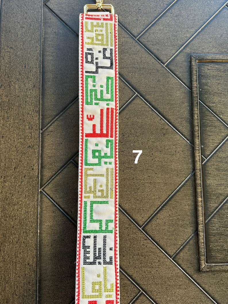 May include: A white strap with red, green, black, and gold embroidered Arabic text. The strap has a gold metal buckle.