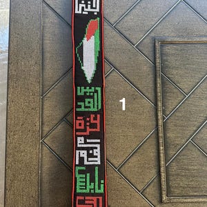 May include: A black fabric strap with a Palestinian flag design and Arabic text. The strap has a silver hook at the top.