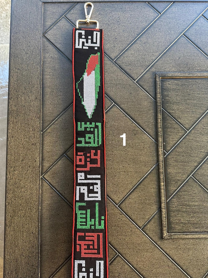 May include: A black fabric strap with a Palestinian flag design and Arabic text. The strap has a silver hook at the top.