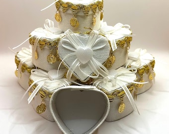 May include: A stack of five white heart-shaped boxes with gold trim and gold coin accents. The boxes are arranged in a pyramid shape, with the largest box at the bottom and the smallest box at the top.