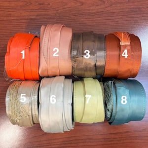 May include: An assortment of eight rolled ribbons in various colors. The colors include orange, light brown, dark brown, rust, beige with fringe, light gray, light green, and teal. The ribbons are neatly rolled and displayed.