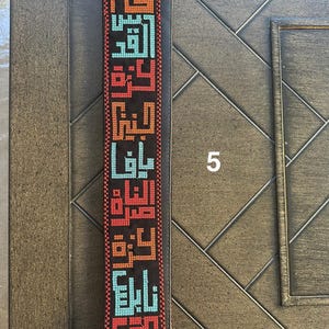 May include: A black strap with colorful Arabic calligraphy embroidered on it. The strap is made of a woven fabric and has a smooth texture.