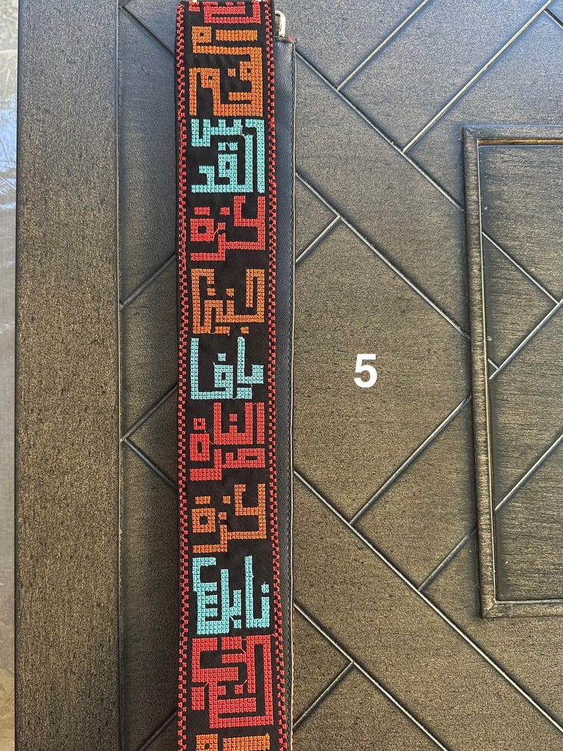 May include: A black strap with colorful Arabic calligraphy embroidered on it. The strap is made of a woven fabric and has a smooth texture.