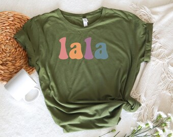 May include: An olive green short-sleeve t-shirt featuring the word lala printed across the chest in a wavy, multi-colored font. Each letter is a different color, including pink, orange, purple, and blue, set against the solid green fabric.