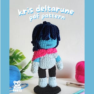 Puede incluir: Muñeca de ganchillo hecha a mano de Kris de Deltarune, con cara y pelo azules, una bufanda rosa y un atuendo azul claro y gris. La muñeca está sobre una base negra. El texto "Kris Deltarune pdf pattern" está en la parte superior.