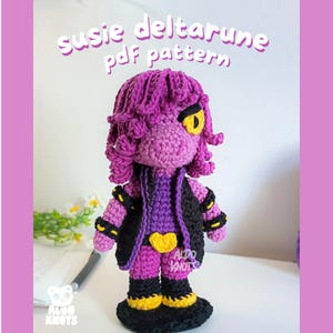 Puede incluir: Muñeca de ganchillo de Susie de Deltarune, principalmente morada con detalles negros. Tiene un chaleco negro, una hebilla en forma de corazón amarilla y ojos amarillos. El texto "susie deltarune pdf pattern" es visible.