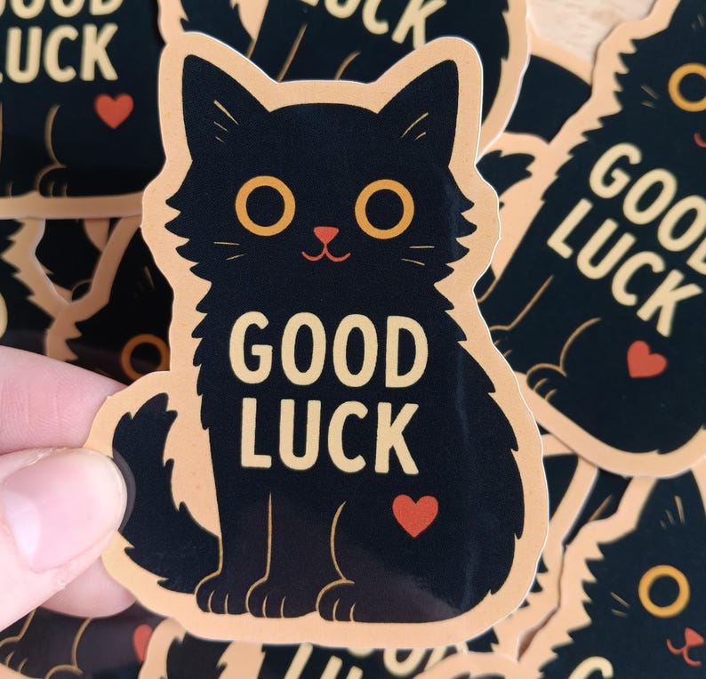 May include: A black cat sticker with a peach background. The cat is sitting with its paws tucked under its body. The text "GOOD LUCK" is written in yellow on the sticker. A small red heart is on the cat's chest.
