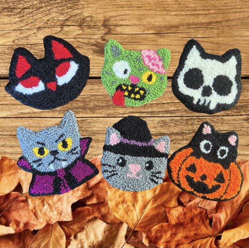 May include: Six colorful, handmade Halloween cat-themed decorations. The designs include a black cat, a zombie cat, a skull cat, a vampire cat, a witch cat, and a jack-o'-lantern cat. The decorations are placed on a wooden surface with autumn leaves.