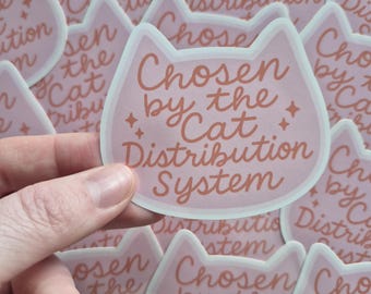 Puede incluir: Una pegatina rosa claro con forma de cabeza de gato con el texto "Chosen by the Cat Distribution System" en cursiva. La pegatina tiene un borde blanco y pequeños detalles de estrellas. Otras pegatinas son visibles en el fondo.