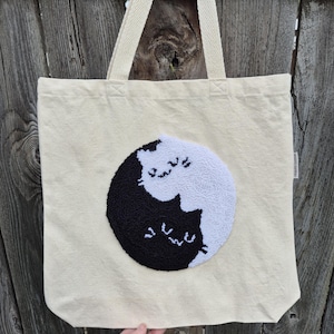 Op de afbeelding: Crèmekleurige draagtas met een zwart-wit kattenontwerp. Het ontwerp toont twee katten in een yin-yang formatie. De tas heeft twee crèmekleurige banden en is gemaakt van een stevige stof. De achtergrond is een verweerde houten schutting.
