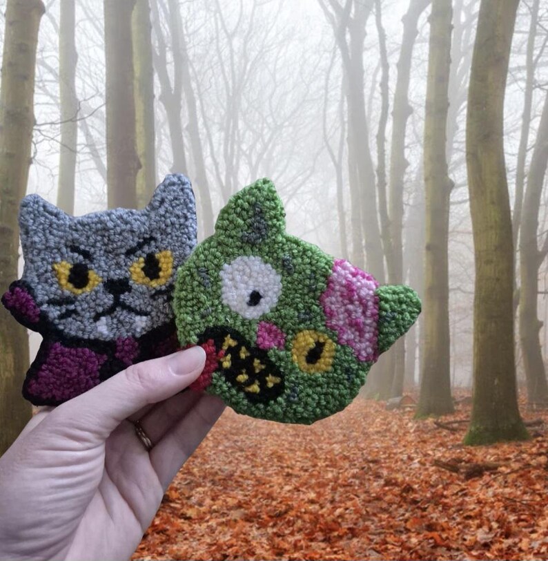 May include: Two handmade cat-shaped and zombie-cat-shaped decorative items. The cat is gray with yellow eyes and a purple collar. The zombie cat is green with pink and yellow accents. The background is a forest with fallen leaves.