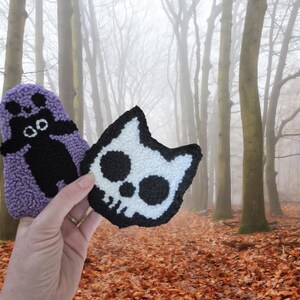 May include: Two handmade, textured patches. One is a purple ghost with a black face and body. The other is a white cat skull with black details and a black border. The background is a forest with fallen leaves.