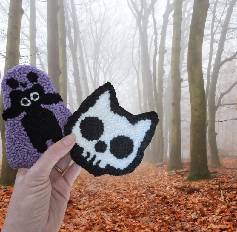 May include: Two handmade, textured patches. One is a purple ghost with a black face and body. The other is a white cat skull with black details and a black border. The background is a forest with fallen leaves.