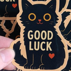 May include: A black cat sticker with a peach background. The cat has yellow eyes and a red heart on its belly. The text "GOOD LUCK" is written in yellow letters on the cat's belly.
