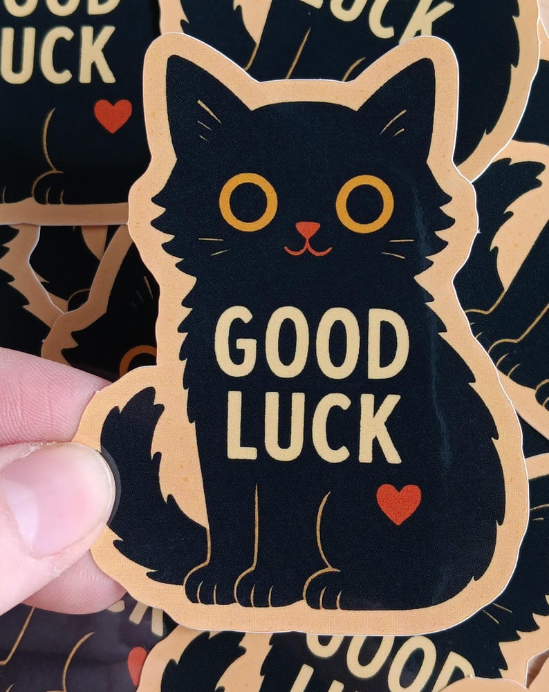 May include: A black cat sticker with a peach background. The cat has yellow eyes and a red heart on its belly. The text "GOOD LUCK" is written in yellow letters on the cat's belly.
