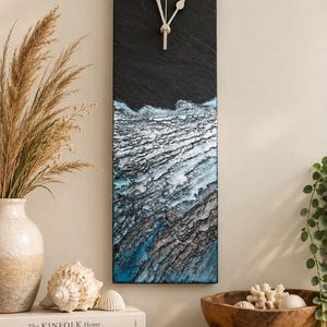 May include: A rectangular wall clock made of black slate with a textured, layered surface featuring blue and white accents that resemble ocean waves. The clock has two simple, light-coloured hands positioned in the upper section of the slate panel.