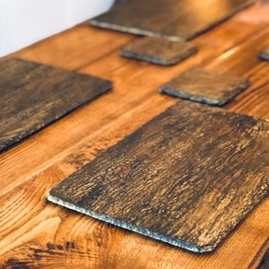 May include: Set of six slate coasters with a rustic wood grain pattern. The coasters are rectangular and have a dark brown colour.