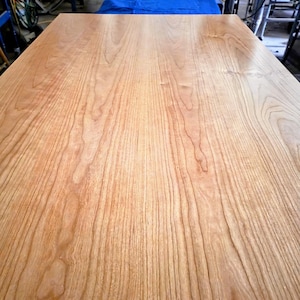 May include: A large, rectangular wooden tabletop with a light brown finish. The wood grain is prominently displayed, featuring vertical lines and subtle variations in color. The surface appears smooth and ready for use.