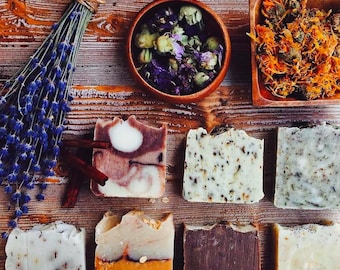 May include: Assortment of eight handmade soap bars with various colors, patterns, and ingredients. The soaps are arranged on a wooden surface with dried lavender, dried flowers, and a bowl of dried herbs.