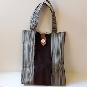 May include: A vertical tote bag with a striped pattern in shades of blue, gray, and beige. The center panel is a solid dark brown. The bag has two handles and a button closure. The bag is approximately 15 inches tall.