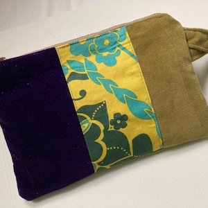 Puede incluir: Un bolso de mano rectangular con un diseño de bloques de color. El lado izquierdo es de terciopelo morado oscuro, el centro presenta un estampado floral amarillo y turquesa, y el lado derecho es de ante verde oliva apagado. Una cremallera dorada y una pequeña correa verde oliva completan el diseño.