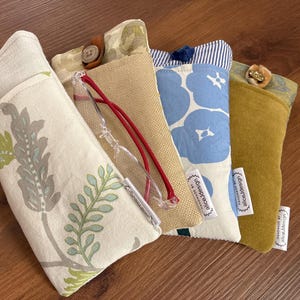 May include: Four rectangular fabric pouches for glasses, each featuring a button closure and a small white tag labeled handmade by alicejdesign. The designs include cream with green leaves, tan, blue with white flowers, and solid mustard yellow.