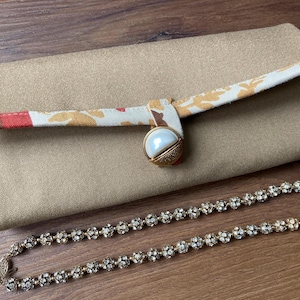 Op de afbeelding: Een beige clutch met een patroonband en een parelachtige sluiting, gecombineerd met een sprankelende strass-ketting. De clutch heeft een bloemenpatroon op de band en een goudkleurige sluiting. De ketting heeft kleine, ronde, sprankelende stenen.