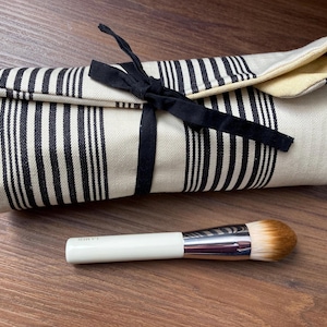 May include: A rolled-up makeup brush roll with black and white stripes, tied with a black ribbon. A white-handled makeup brush with a silver ferrule and brown bristles is next to the roll.